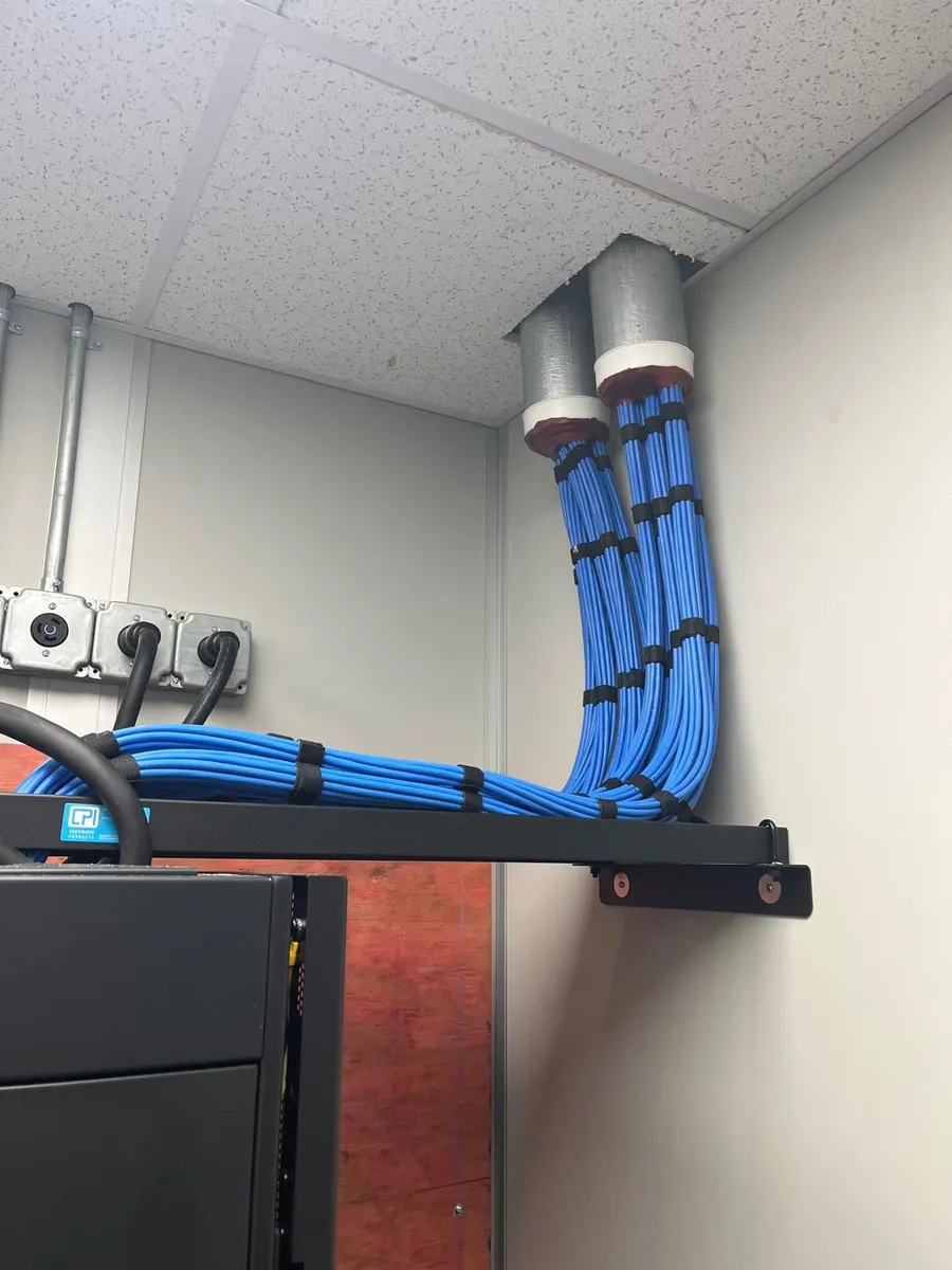 Cable bundles coming through firestopped wall sleeves onto a ladder rack above a network cabinet